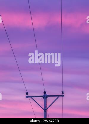 Dieses Bild zeigt die Silhouette eines elektrischen Pylons vor einem atemberaubenden Sonnenaufgangshimmel in lila und rosa Tönen. Die Stromleitungen s Stockfoto