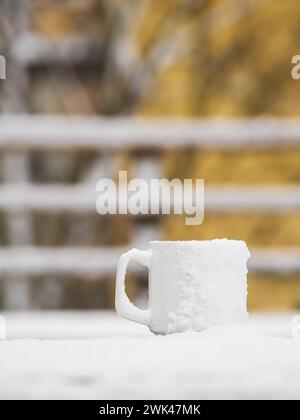 In dieser ruhigen Szene sitzt auf einem Holzgeländer ein weißer Becher mit frischem Schnee, der einen starken Kontrast zu den gelben Herbstblättern bildet, die A sind Stockfoto