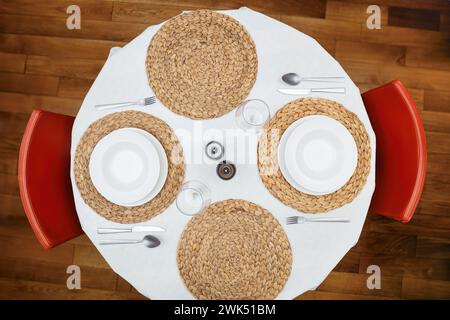 Blick von oben auf einen runden Esstisch mit weißen Tellern, Besteck, gewebten Tischsets und roten Stühlen, bereit für eine Mahlzeit. Stockfoto