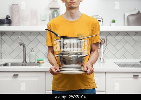 Männliche Balancierung mit einem Haufen dreckiger und gebrauchter Küchenutensilien, Teller, Pfannen im modernen Küchenhintergrund, Haushaltsreinigung Stockfoto