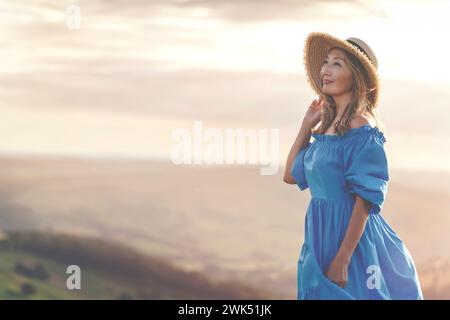 asiatische Frau in einem blauen Kleid und einem Hut mit einem Lavendelstrauß in den Händen auf einem Gipfel des Berges bei Sonnenuntergang im Sommer Stockfoto