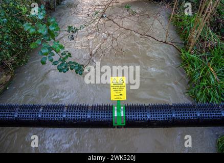 Rohr über Fluss mit Kletterstacheln, die mit gelbem Warnschild umschnallt sind. Stockfoto