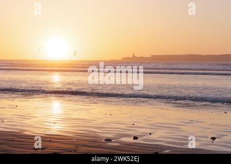 Sonnenuntergänge über einem Sandstrand mit Kitesurfern und einer Insel am Horizont in Essaouira, Marokko. Februar 2024 Stockfoto