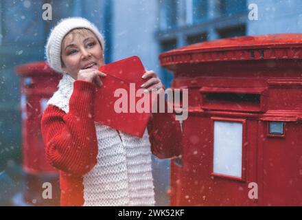 Frau in rotem Mantel und weißem Hut, die eine Karte in den roten Briefkasten steckt und an einem schneebedeckten Tag durch eine englische Stadt spaziert Stockfoto