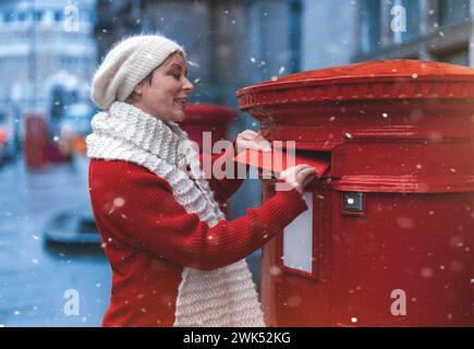 Frau in rotem Mantel, die eine Karte in den roten Briefkasten steckt und an einem schneebedeckten Tag durch eine englische Stadt läuft Stockfoto
