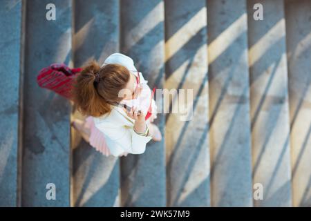 Obere Ansicht einer eleganten Frau mittleren Alters in rosa Kleid und weißer Jacke in der Stadt mit roter Tasche und Sonnenbrille, die auf einem Smartphone spricht. Stockfoto