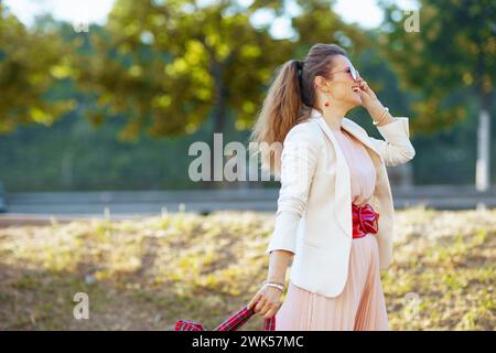 Glückliche trendige Frau in rosa Kleid und weißer Jacke in der Stadt mit Sonnenbrille zu Fuß. Stockfoto