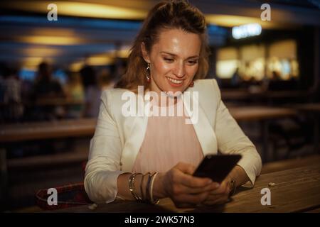 Glückliche trendige Frau in rosa Kleid und weißer Jacke in der Stadt mit Smartphone-App im Straßencafé. Stockfoto