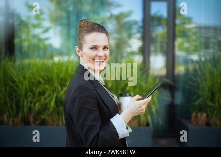 Glückliche moderne Geschäftsfrau in der Nähe des Bürogebäudes in schwarzer Jacke mit einer Tasse Kaffee über Smartphone. Stockfoto