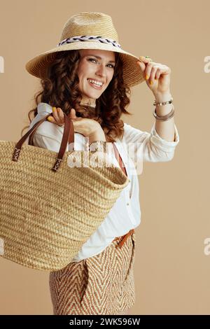 Strandurlaub. Lächelnde elegante 40 Jahre alte Hausfrau in weißer Bluse und Shorts vor beigefarbenem Hintergrund mit Strohbeutel und Strohhut. Stockfoto