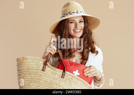 Strandurlaub. Glückliche trendige 40-jährige Frau in weißer Bluse und Shorts isoliert auf Beige mit Verbandskasten, Strohtasche und Sommerhut. Stockfoto