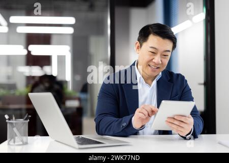 Lächelnder und verschlafener junger asiatischer Geschäftsmann, Geschäftsführer sitzt im Büro, arbeitet an einem Laptop und benutzt ein Tablet. Stockfoto