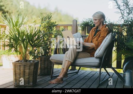 Frau, die im Garten arbeitet, mit Laptop auf den Beinen. Geschäftsfrau, die aus der Ferne vom Heimbüro im Freien arbeitet, leichtes Mittagessen, Sandwich isst. Konzept von Out Stockfoto