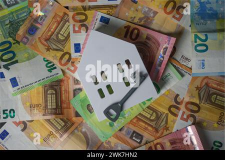 Ein großer Haufen Euro-Geld und ein weißes Holzhaus und ein Schlüssel auf schwarzem Hintergrund - von oben. Stockfoto