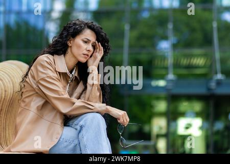 Besorgte junge Frau, die draußen sitzt, eine Brille in der Hand hält, tief in Gedanken mit einem beunruhigten Ausdruck, urbaner Hintergrund. Stockfoto