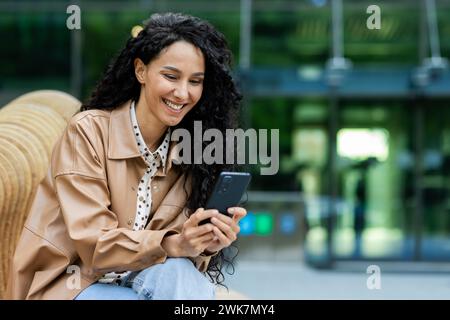 Professionelle junge hispanische Geschäftsfrau, die eine Pause vor einem Bürogebäude genießt und gerne mit ihrem Smartphone interagiert. Stockfoto