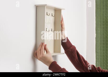 Die junge Frau hängt einen hölzernen Schlüsselhalter an die Wand und zieht in das neue Apartment oder das neue Wohnzimmerkonzept Stockfoto