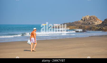 Ein Tourist mit Hut, der am Sandstrand mit Flipflops spaziert. Ein Mann, der an der Küste in Goa Indien entlang läuft. Sandstrände in Goa. Stockfoto