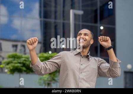 Glücklicher hispanischer junger Mann tanzt in der Nähe von Bürozentrum und Gerichtsgebäude, glücklicher Erfolg, der die Arme hebt. Nahaufnahme. Stockfoto