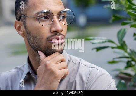 Ein Nahfoto eines jungen hispanischen Mannes, der vor der Stadt mit Brille steht, seinen Bart mit der Hand reibt und ernsthaft und nachdenklich zur Seite blickt. Stockfoto