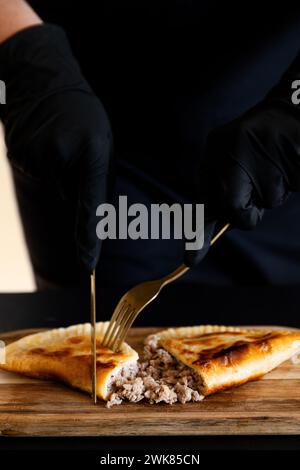 Hände in schwarzen Handschuhen schneiden Cheburek, eine Torte mit Fleisch.vertikalem Rahmen Stockfoto