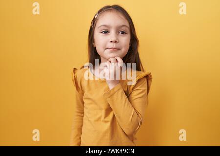 Das süße hispanic Girl steht stolz und selbstbewusst, die Arme sind gekreuzt und Hand auf dem Kinn und strahlt ein positives Lächeln auf die Kamera vor einer hellgelben isola Stockfoto