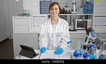 Die leuchtende junge blonde Wissenschaftlerin, die sich mit Notizen auf ihrem Klemmbrett beschäftigt, wirft ein warmes Lächeln über ihr geschäftiges Labor, voll vertieft in Biowissenschaften Stockfoto