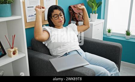 Hispanische Frau, die sich in einem modernen Büro mit einem Laptop entspannt und einen Moment der lockeren Geschäftspause zeigt. Stockfoto
