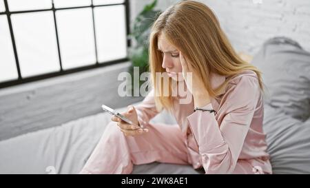Blonde Frau im pinkfarbenen Pyjama, die konzertiert auf das Smartphone in ihrem modernen Schlafzimmer schaut. Stockfoto