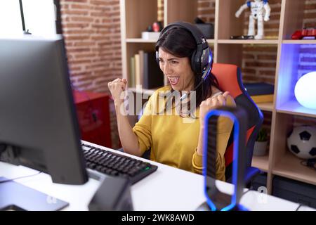 hispanische Frau im mittleren Alter, die Videospiele mit Kopfhörern spielt sehr glücklich und aufgeregt, mit erhobenen Armen, Lächeln und Schreien die Siegergeste zu machen Stockfoto