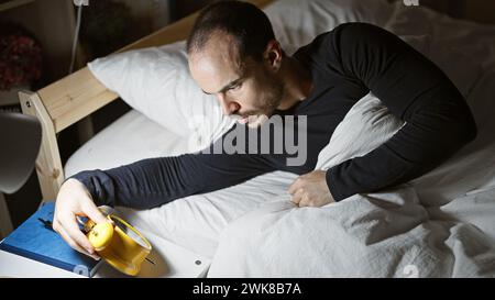Ein kahler Mann mit Bart, der nachts in einem schwach beleuchteten Schlafzimmer einen Wecker auf eine gelbe Uhr stellt. Stockfoto