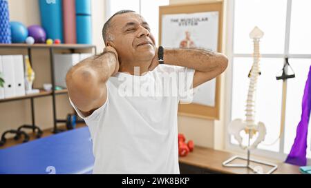 Ein hispanischer Mann mittleren Alters dehnt sich in einer hellen Reha-Klinik aus und zeigt einen Moment der Entspannung und Physiotherapie. Stockfoto