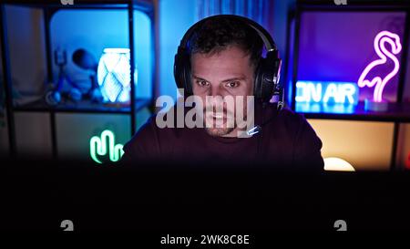 Konzentrierter junger hispanischer Mann mit Bart im Spielzimmer, der nachts Kopfhörer trägt. Stockfoto