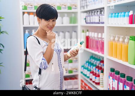 chinesische Kundin im mittleren Alter lächelt selbstbewusst in der Hand einer Shampoo-Flasche in der Apotheke Stockfoto