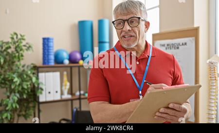 Gutaussehender Seniorenmann mit grauem Haar in einer Klinik hält ein Klemmbrett, das eine professionelle Reha-Umgebung widerspiegelt. Stockfoto