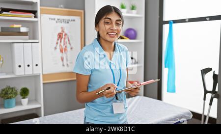 Eine lächelnde junge indianerin mit langen Haaren, die in medizinischen Peelings gekleidet ist, steht in einer Klinik mit einem Klemmbrett. Stockfoto