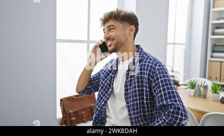 Selbstbewusster junger arabischer Geschäftsmann, der sich während der Arbeit im Büro freudig mit seinem Smartphone unterhalten kann, ein glücklicher Mann steht und über den Erfolg lächelt Stockfoto