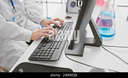 Professionelle Wissenschaftlerin, die auf einer Tastatur in moderner Laborumgebung mit bunten erlenmeyerkolben im Hintergrund tippt. Stockfoto