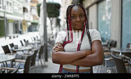 Fröhliche afroamerikanische Frau, die Selbstvertrauen ausdrückt, Arme gekreuzt und strahlend lächelnd in einer zwanglosen Straße Coffee Shop Terrasse unter sonniger urbaner Natur Stockfoto