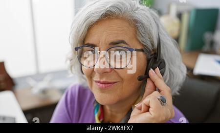 Eine grauhaarige Frau mit Brille trägt in einem modernen Büro ein Headset, das die Professionalität des Kundendienstes darstellt. Stockfoto