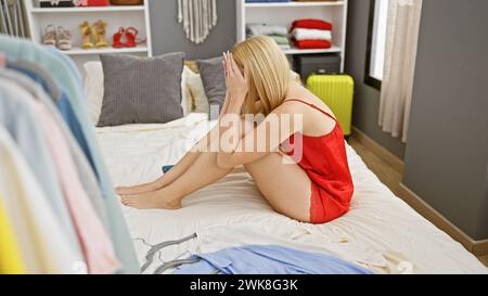 Verärgerte blonde Frau in Rot, die auf dem Bett sitzt, mit verstreuten Klamotten, die Stress vermitteln. Stockfoto