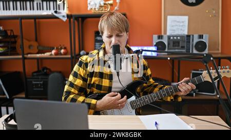Ein junger, gutaussehender, bärtiger Mann singt in ein Mikrofon, während er in einem orangen Musikstudio eine schwarze Gitarre spielt. Stockfoto