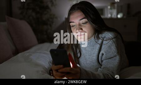 Hispanische Frau mit Smartphone in dunklem Schlafzimmer nachts. Stockfoto