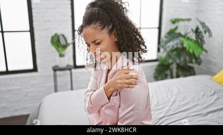Junge, schöne hispanische Frau im Morgenbett, unter starken Schulterschmerzen, unglücklich an den schmerzenden Bereich im Schlafzimmer Stockfoto