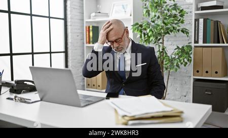 Mann im Anzug mit Glatze, gestresst am Schreibtisch mit Laptop in heller Büroumgebung Stockfoto