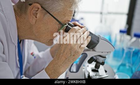 Engagierte grauhaarige Wissenschaftlerin tauchte in intensive Mikroskopforschung auf phd-Niveau im Labor ein Stockfoto