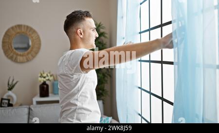 Ein junger hispanischer Mann passt die Vorhänge in einem hell beleuchteten, stilvollen Wohnzimmer an und erweckt so ein Gefühl von Zuhause und Entspannung. Stockfoto