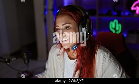 Fesselnder Live-Stream, strahlende junge Rothaarige Streamerin, lächelnd, High-Tech-Gaming im gemütlichen Raum, Kopfhörer an Stockfoto