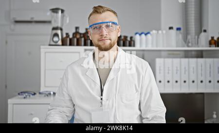 Ein junger bärtiger Mann mit Schutzbrille und Labormantel posiert selbstbewusst in einem klinischen Labor. Stockfoto