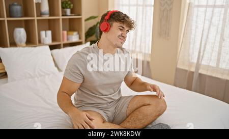 Attraktiver junger hispanischer Mann, der bequem seinen Hals ausstreckt, in seine morgendliche Musik vertieft, auf dem Bett sitzt, Kopfhörer auf, in seinem C Stockfoto